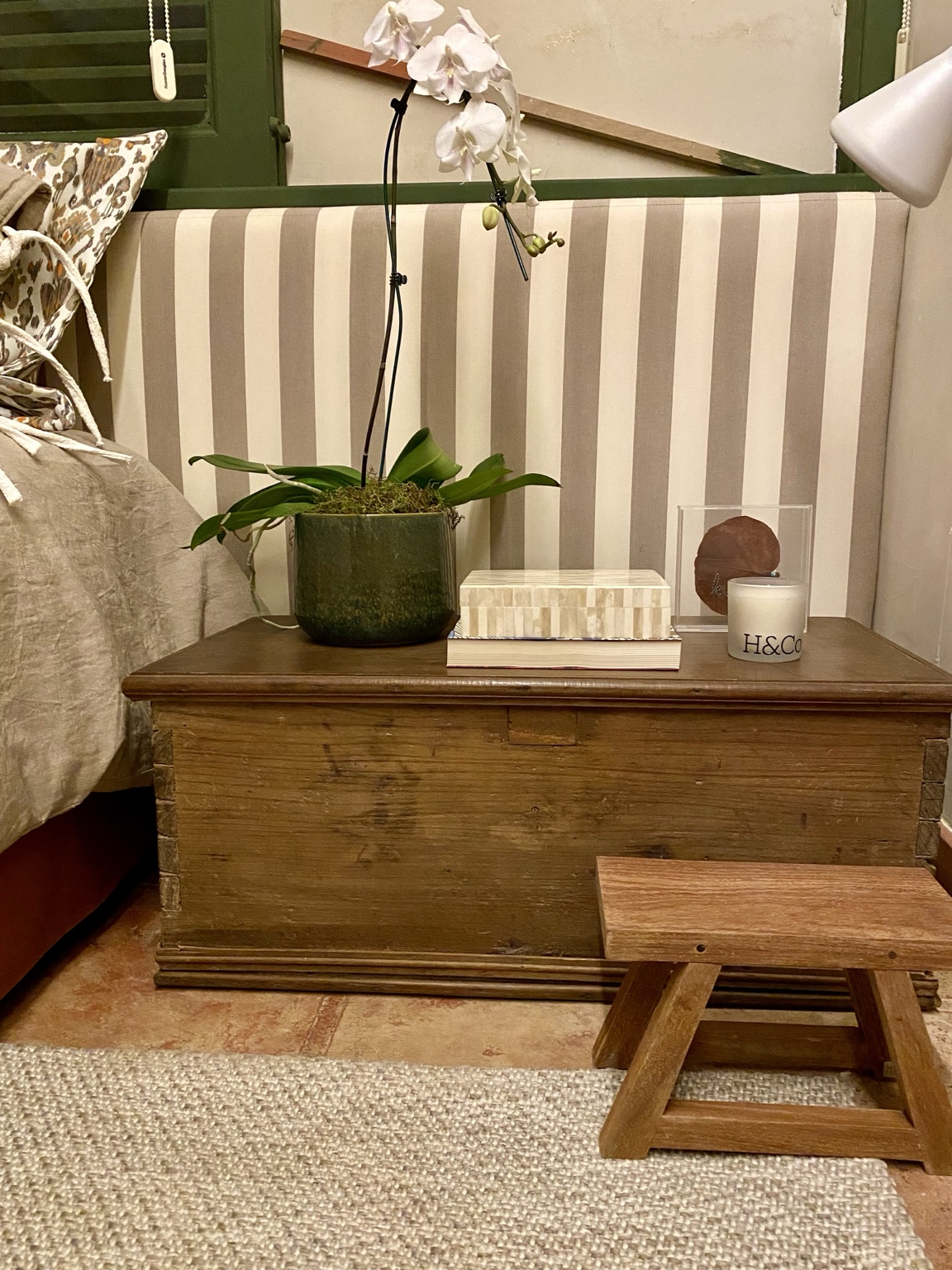 Vela Hyooga & Co. em pote branco sobre baú de madeira ao lado da cama, com orquídea e livros