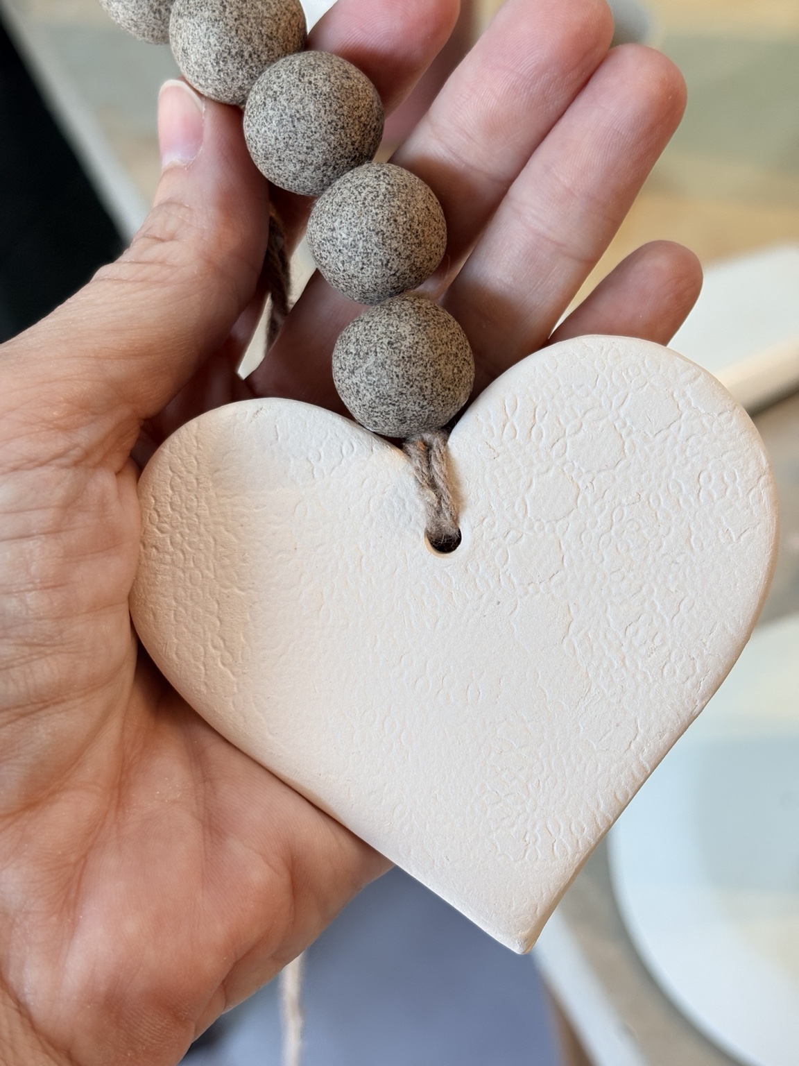 Scent Heart de cerâmica branca com textura artesanal e contas em tom de pedra, apoiado sobre uma mão