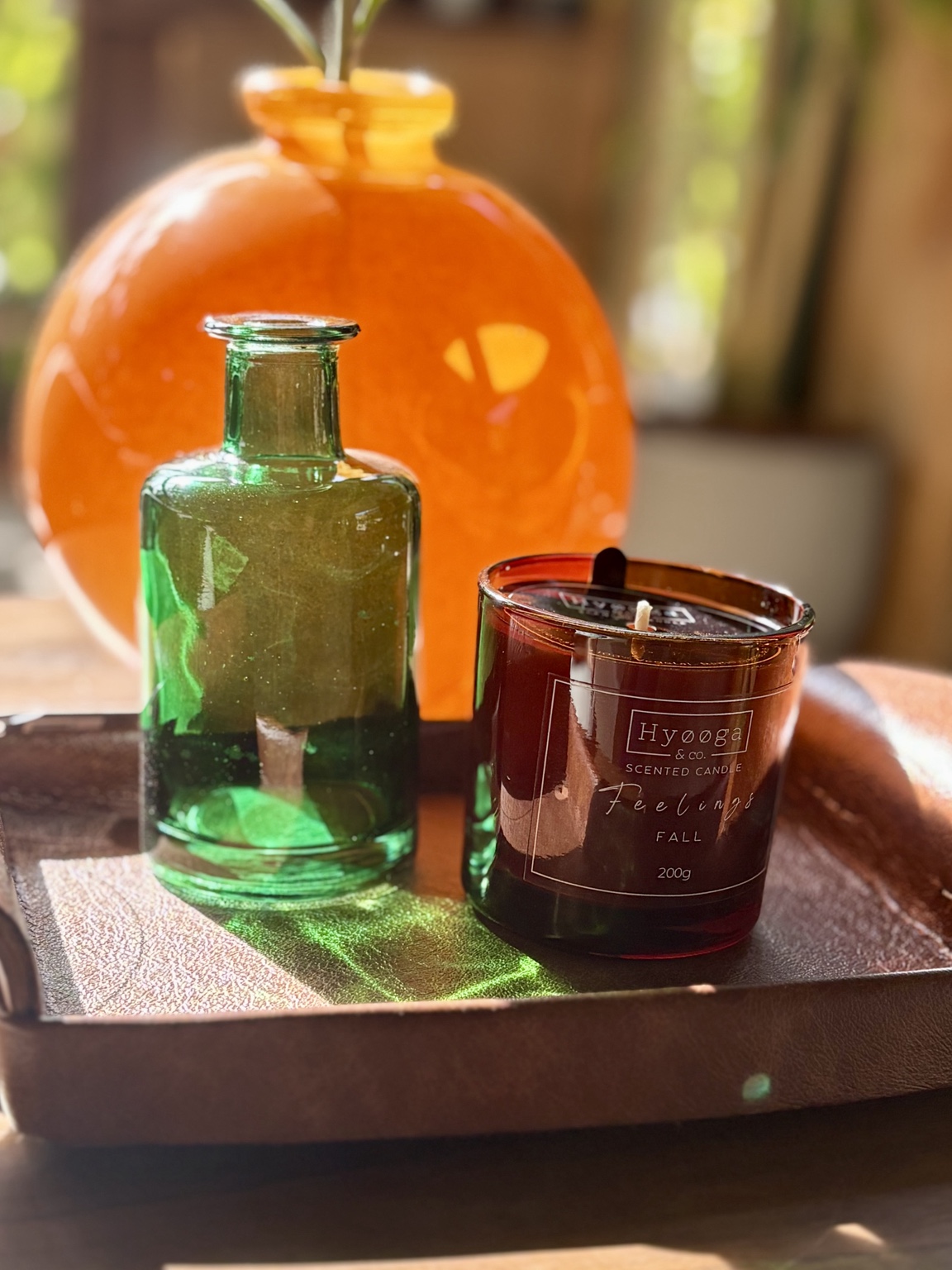 Vela Hyooga & Co. em copo âmbar ao lado de garrafa de vidro verde sobre bandeja de madeira, com vaso laranja ao fundo