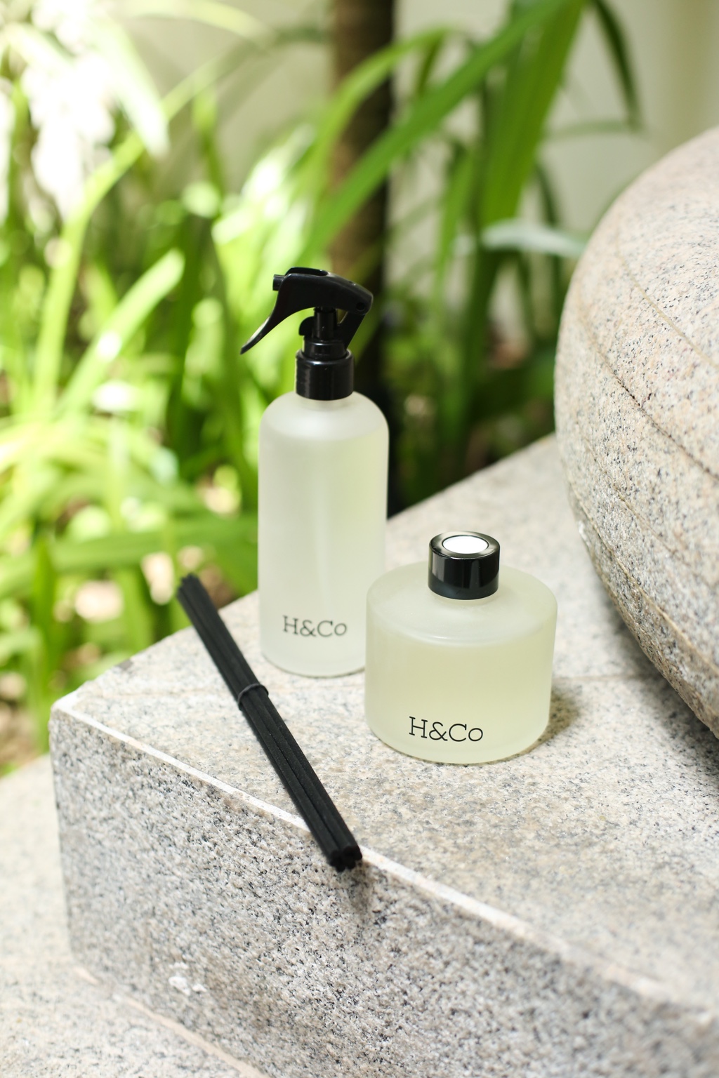 Home spray e difusor Hyooga & Co. em frascos de vidro fosco sobre banco de pedra, com varetas pretas ao lado e folhagem verde ao fundo