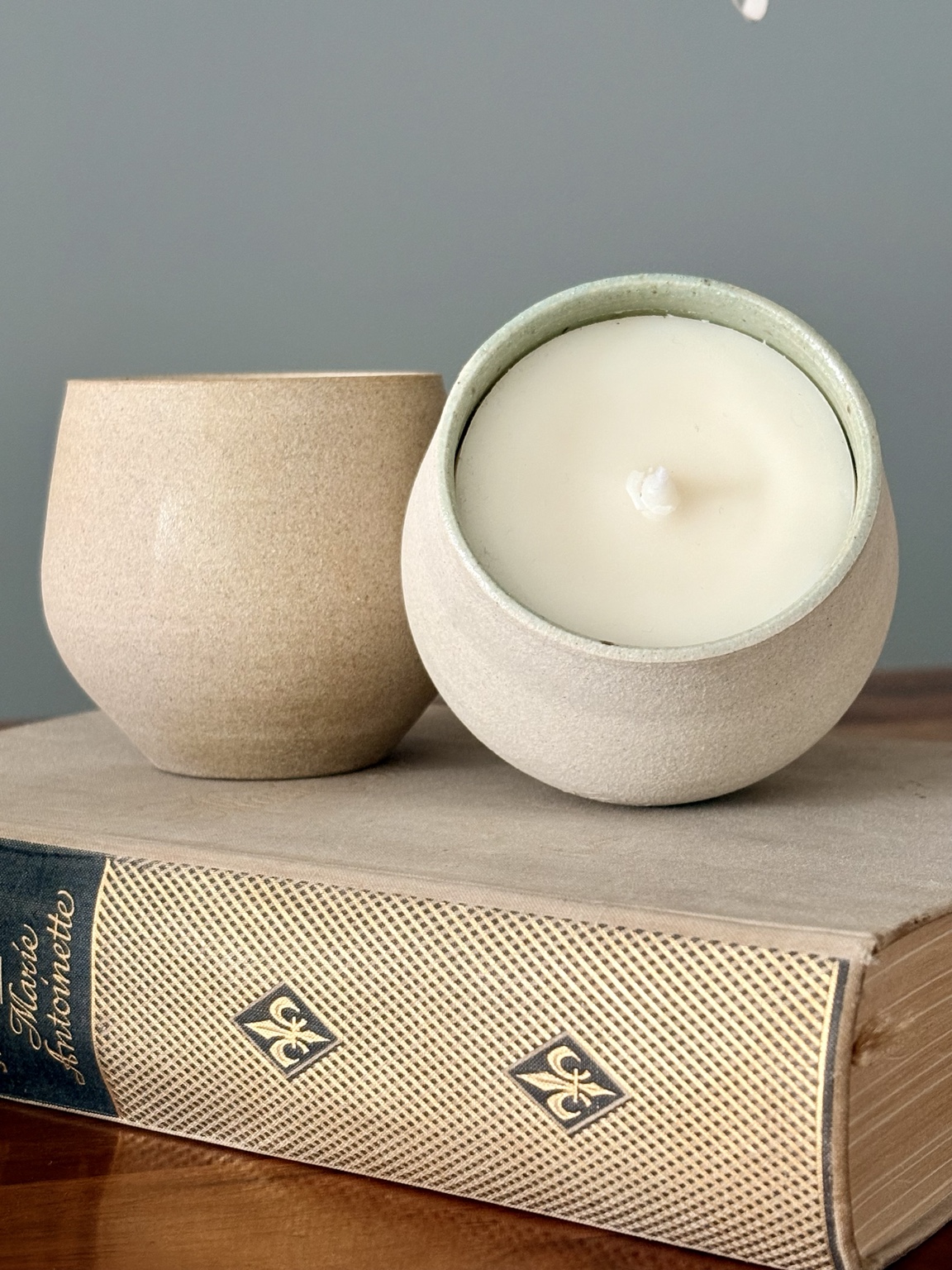 Duas velas Hyooga & Co. em recipientes de cerâmica clara com textura granulada, apoiadas sobre um livro