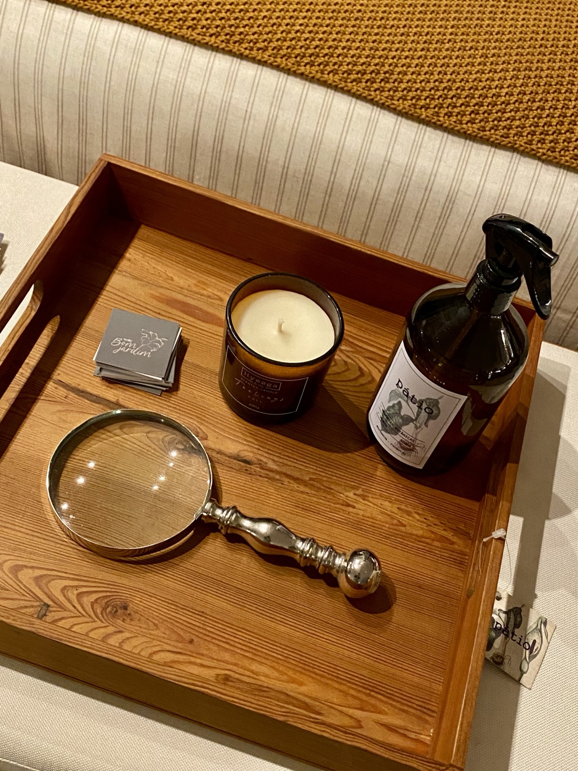 Bandeja de madeira com vela Hyooga & Co., frasco âmbar com borrifador e lupa de metal, sobre sofá claro